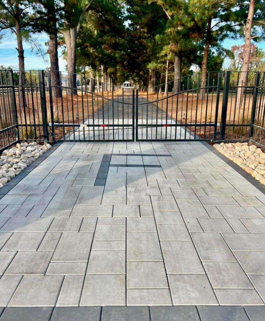 Tree-lined paver driveway with dark inlay pattern leading to a black metal entrance gate at a Virginia property.