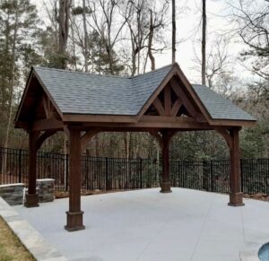 Custom timber-frame pavilion with a gable roof built on a stone paver patio in a wooded Virginia backyard.